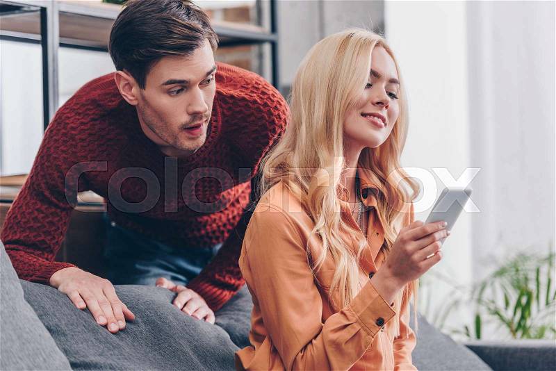 Shocked jealous man looking at smiling young woman using smartphone at home, mistrust concept, stock photo
