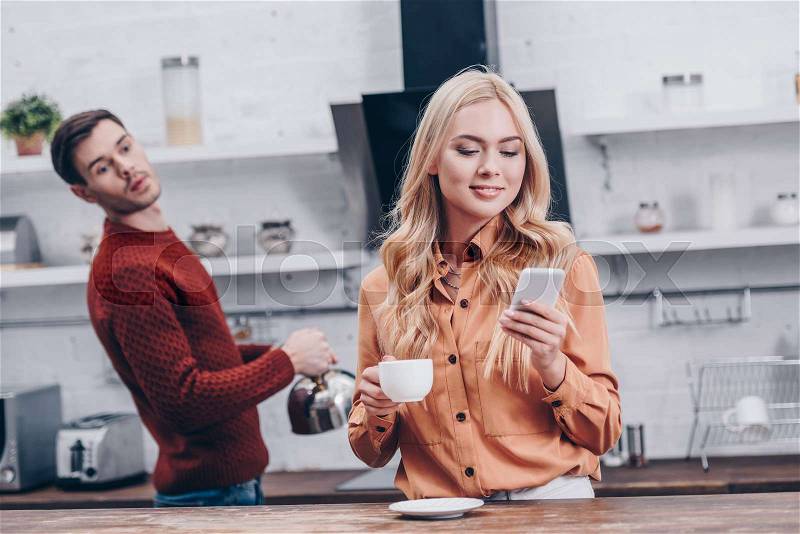 Jealous young man looking at smiling girlfriend holding cup and using smartphone in kitchen, stock photo