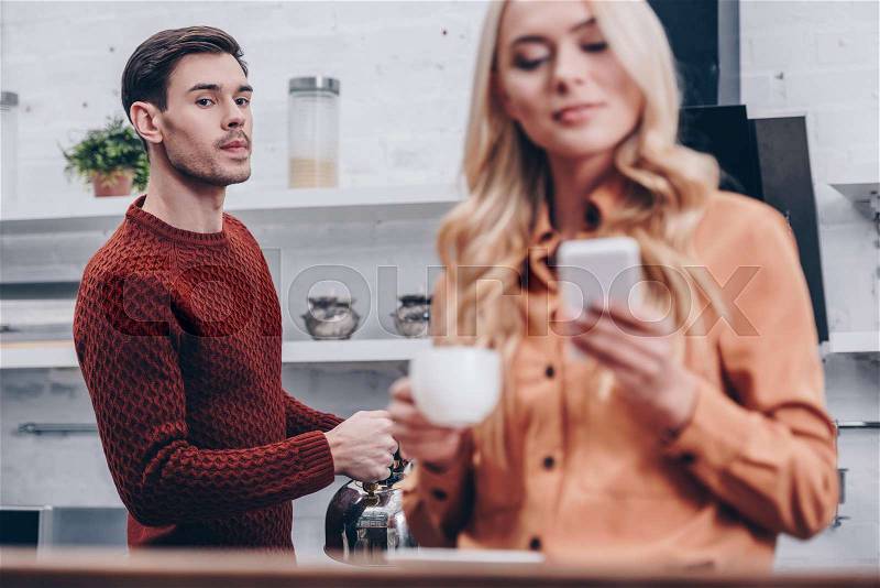 Selective focus of jealous man looking at smiling girlfriend holding cup and using smartphone in kitchen, stock photo