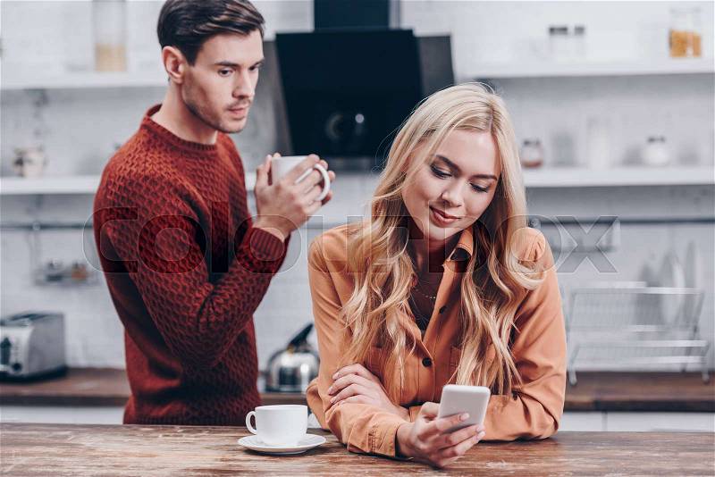 Jealous young man looking at smiling girlfriend using smartphone in kitchen, distrust concept, stock photo