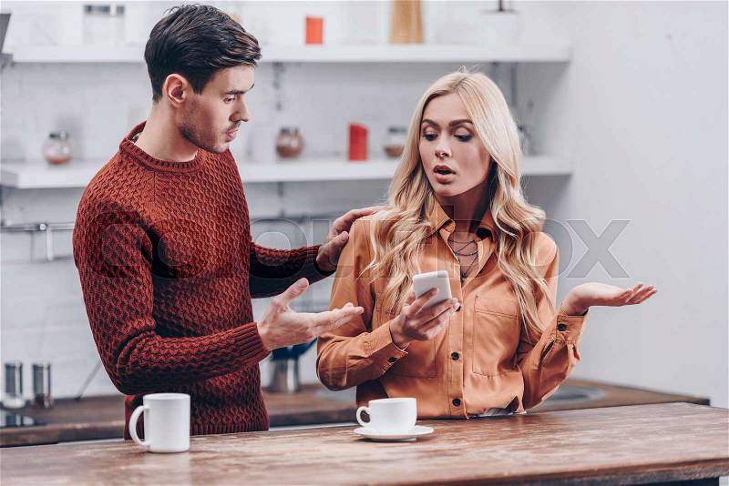 Jealous young man looking at emotional girlfriend using smartphone in kitchen, distrust concept, stock photo