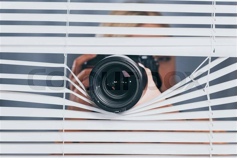 Young woman photographing with camera and spying through blinds, stock photo