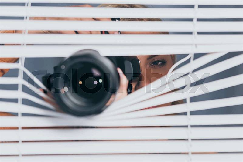 Close-up view of camera and young woman taking pictures and spying through blinds , stock photo