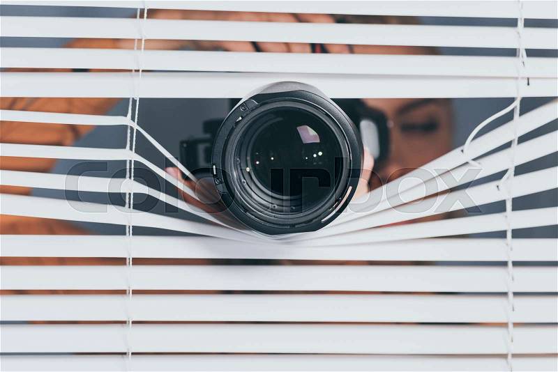 Close-up view of camera and young woman spying through blinds , stock photo