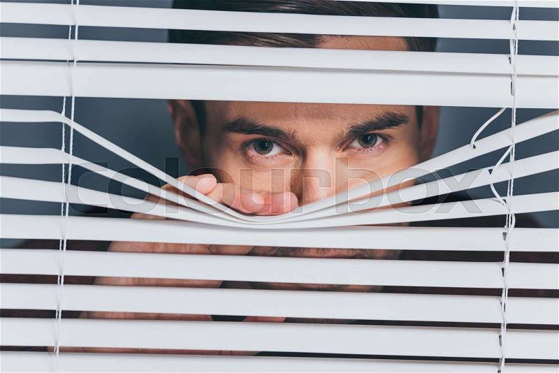 Suspicious young man looking at camera through blinds, mistrust concept, stock photo