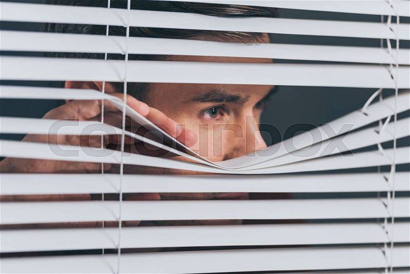 Close-up view of young man spying and looking away through blinds, stock photo