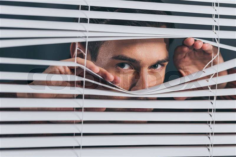 Young man spying and looking at camera through blinds, stock photo