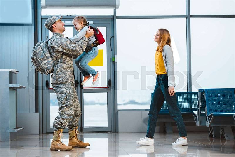 Happy father in military uniform holding in arms cute daughter near wife in airport , stock photo