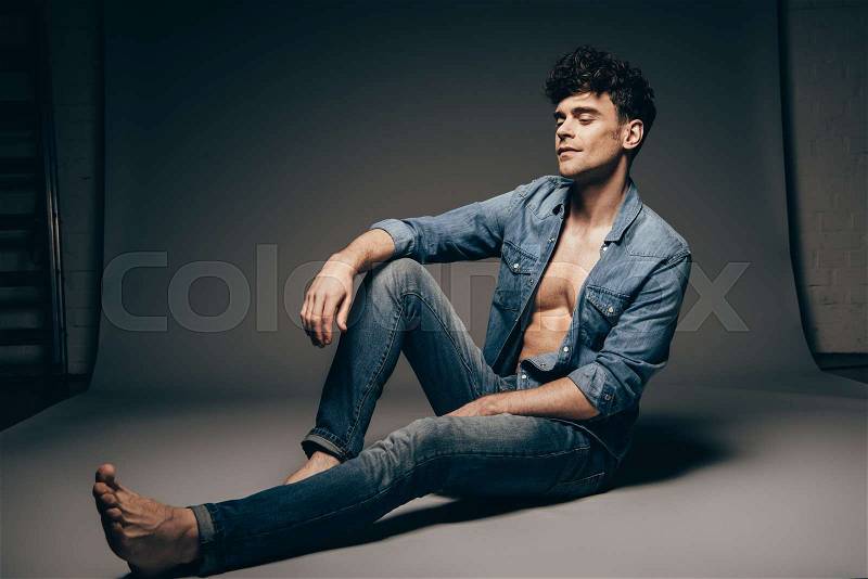 Handsome barefoot man sitting in jeans clothes on dark grey, stock photo