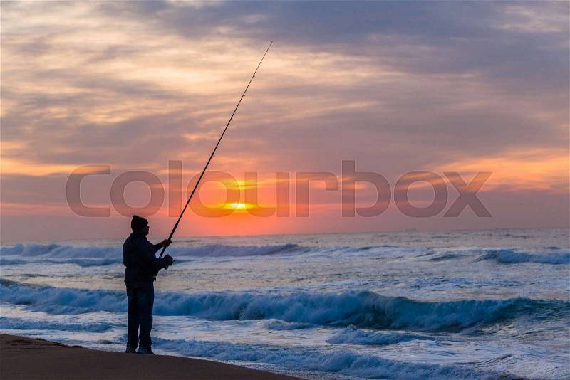 Fisherman fishing silhouetted unidentified on beach waters edge with ocean waves early morning dawn sunrise, stock photo