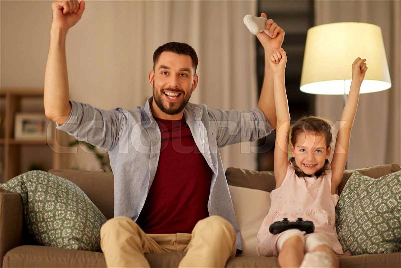 Family, gaming and entertainment concept - happy father and little daughter with gamepads celebrating triumph in video game playing at home in evening, stock photo
