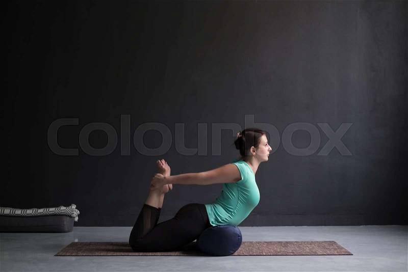 Young woman practicing yoga Bow pose, Dhanurasana using bolster. Therapy variation. Side view, stock photo