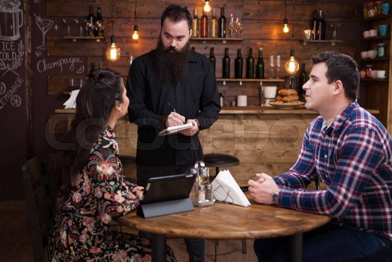 Friendly waiter is taking order from beautiful young couple. Vintage pub, stock photo