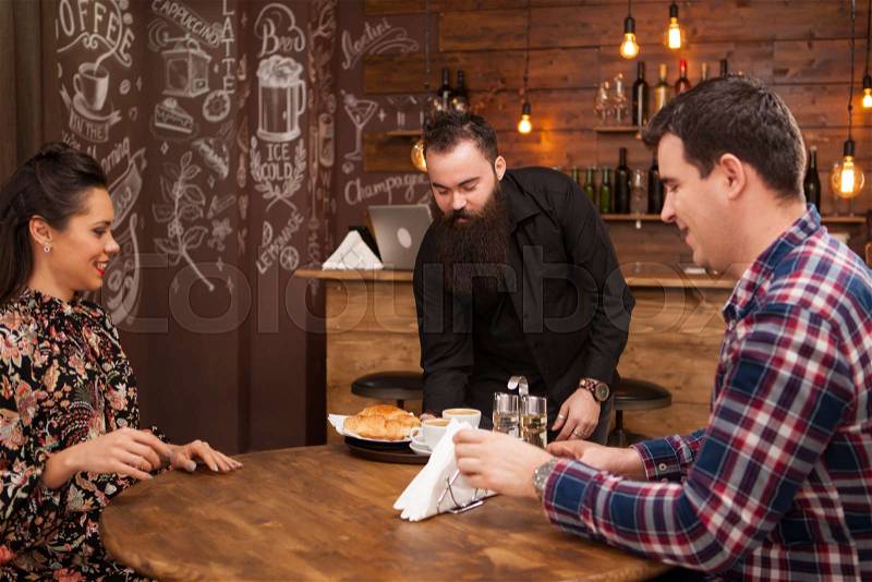 Young waiter serving food to male and female customers at table in cafe. Hipster pub, stock photo