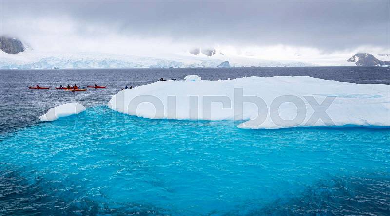 Huge iceberg with blue underwater part and small tourists kayaks with antarctic lagoon in the backgound, Peterman island, Antarctic peninsula, stock photo