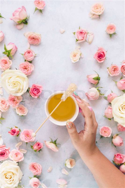 Flat lay with woman\'s hand holding cup of tea surrounded by various roses and petals, textured background. Morning, drink, break, stock photo