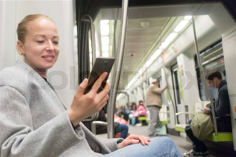 Beautiful cheerful blonde caucasian woman wearing winter coat and jeans using smart phone while traveling by metro. Public transportation concept, stock photo