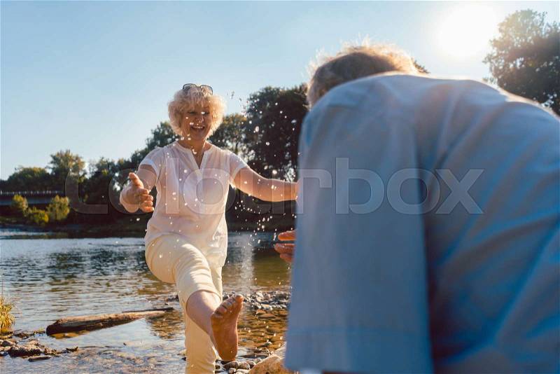 Full length of a funny senior couple playing with water at the river while enjoying their happy relationship in a sunny day of summer, stock photo