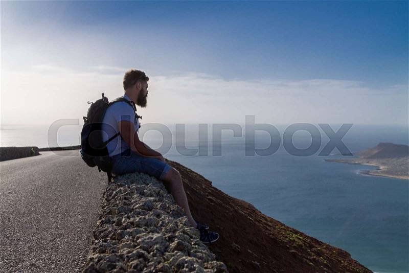 A guy in summer clothes is sitting on a stone fence and looks at a nearby island in the ocean. Mirador del Rio, Lanzarote, Spain, stock photo