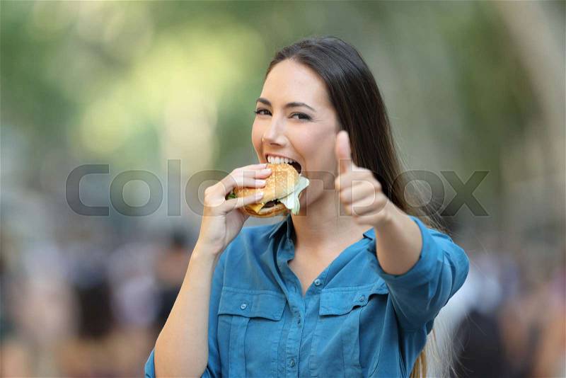 Happy woman eating a burger gesturing thumb up in the street, stock photo