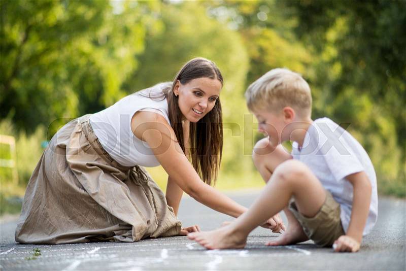 A small boy with mother drawing hopscotch with chalk on a road in park on a summer day, stock photo