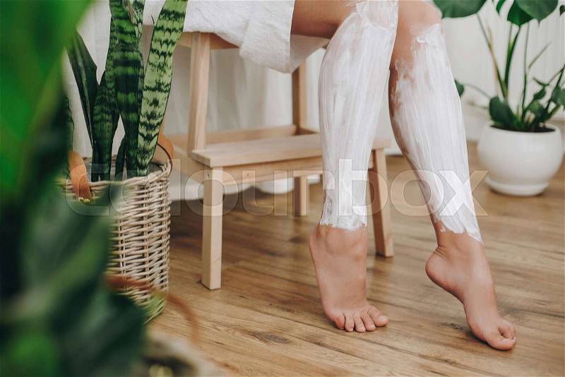 Hair Removal concept, depilation cream. Young woman in white towel applying shaving cream on her legs in home bathroom with green plants. Skin care and wellness ..., stock photo