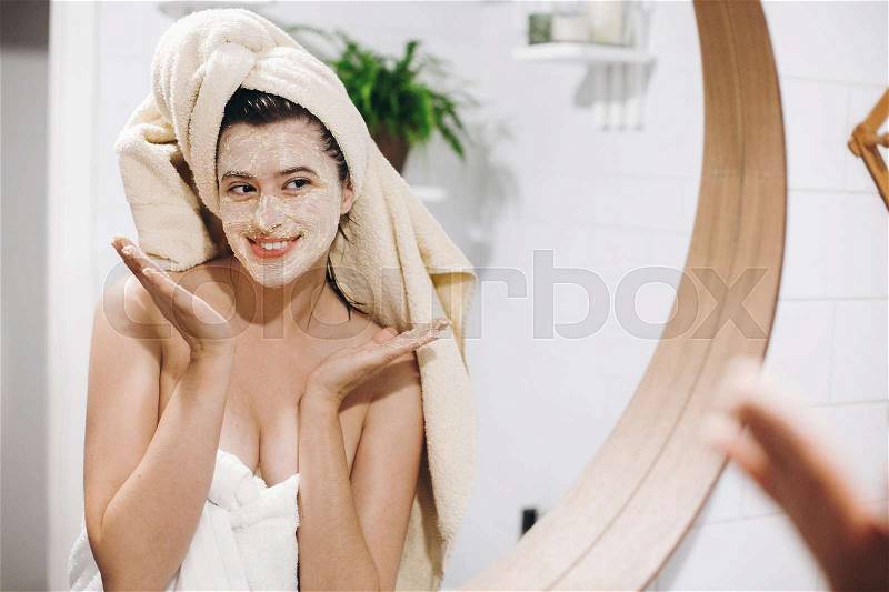 Young happy woman in towel applying organic face scrub and looking at round mirror in stylish bathroom. Girl making facial massage, peeling and cleaning skin on ..., stock photo