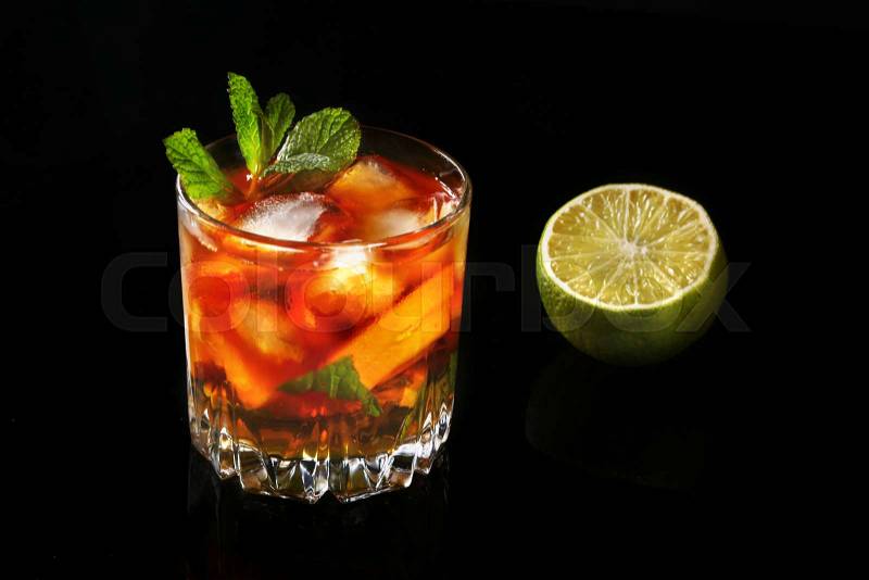 Glass of Dark Rum Cocktail with lime, orange, ice cubes and mint leaves on black mirror background, stock photo