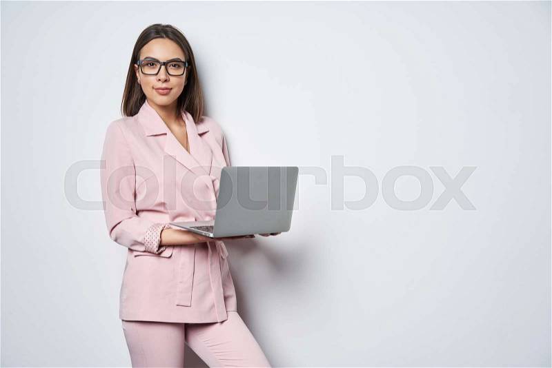 Confident business woman wearing pink suit standing by white wall holding opened laptop looking at camera, stock photo