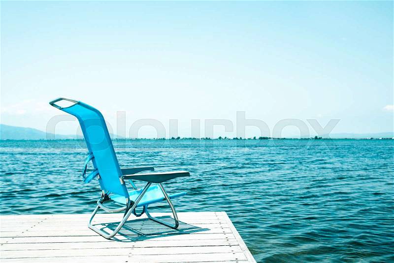 A lonely blue deck chair at the end of a weathered wooden pier next to the water, in a relaxing ambient, with a blank space on the clear sky, stock photo