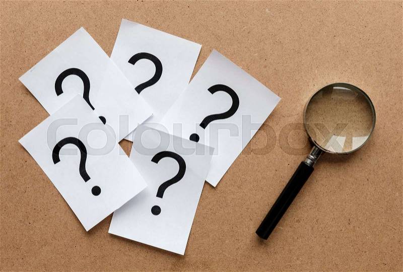 Question marks printed on white cards lying on a wooden background with a magnifying glass in a conceptual image, stock photo