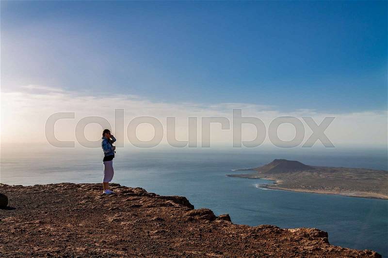 A woman stands on the edge of a cliff and looks at a nearby island in the ocean. Wind ruffles hair. Mirador del Rio, Lanzarote, Spain, stock photo