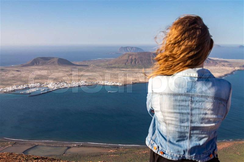 A woman stands on the edge of a cliff and looks at a nearby island in the ocean. Wind ruffles hair. Mirador del Rio, Lanzarote, Spain, stock photo
