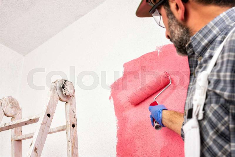 Caucasian house painter worker in white work overalls, with the roller he paints the wall with the colored painting of pink. Construction industry. Bottom view, stock photo