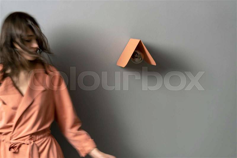 Blurred woman with windy hair in a coral trench near a metal colorful lamp with an edison light bulb on the gray wall background indoors. Horizontal, stock photo