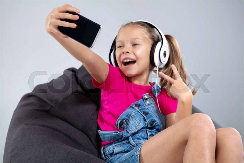 Happy little schoolgirl using smartphone with headphones making selfie. Children and technology concept, stock photo
