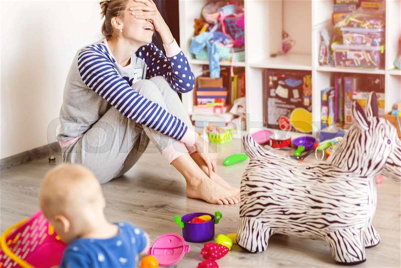 Tired of everyday household mother sitting on floor with hands on face. Kid playing in messy room. Scaterred toys and disorder. Happy parenting, stock photo