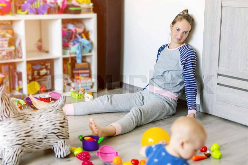 Tired of everyday household mother sitting on floor with hands on face. Kid playing in messy room. Scaterred toys and disorder. Happy parenting, stock photo