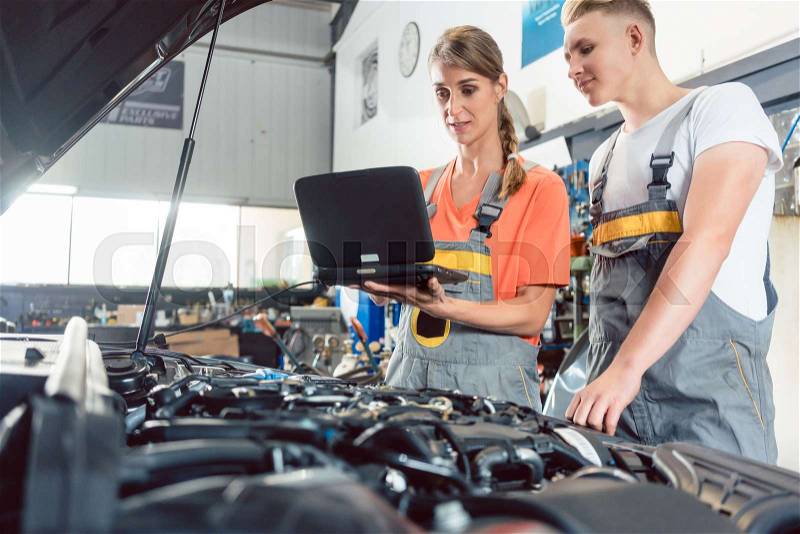Experienced female auto mechanic checking the engine error codes scanned by a car diagnostic software while standing next to a motivated apprentice, stock photo