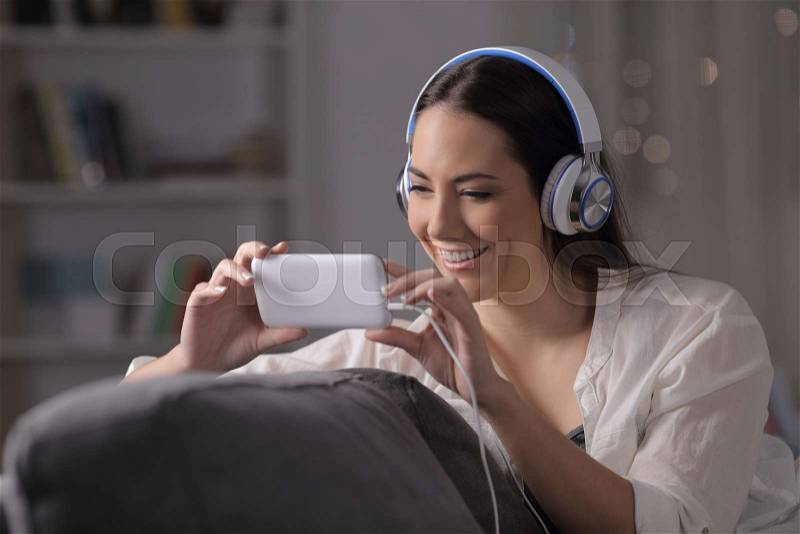 Happy woman watches videos wearing headphones sitting on a couch in the night at home, stock photo