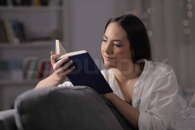 Serious lady reads a book sitting on a couch in the night at home, stock photo
