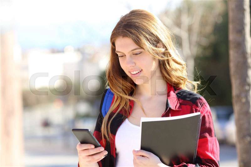 Relaxed student is using a smart phone walking in the street a sunny day, stock photo