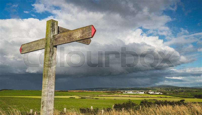 Close-up cross road signpost on the rainy sky background. Northern Ireland landscape. The wooden guidepost with the red arrows. The green grass covered fields before ..., stock photo