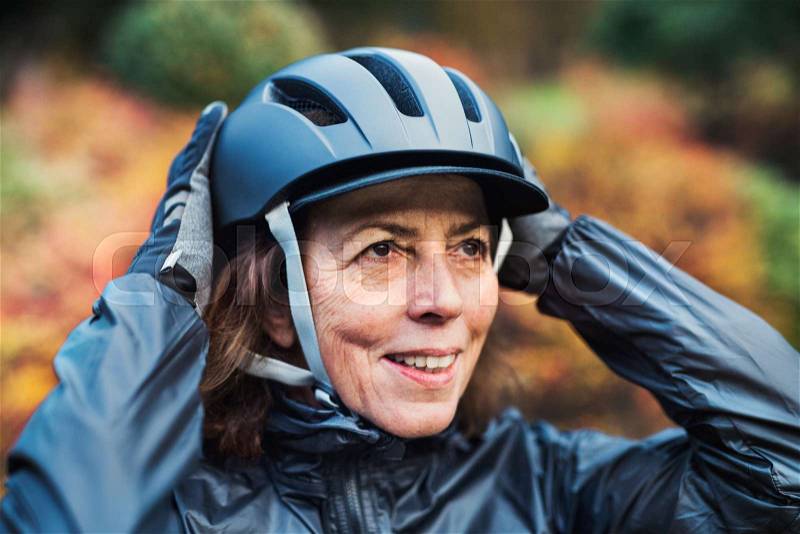 A close-up of active senior woman standing outdoors in nature, putting on bicycle helmet, stock photo