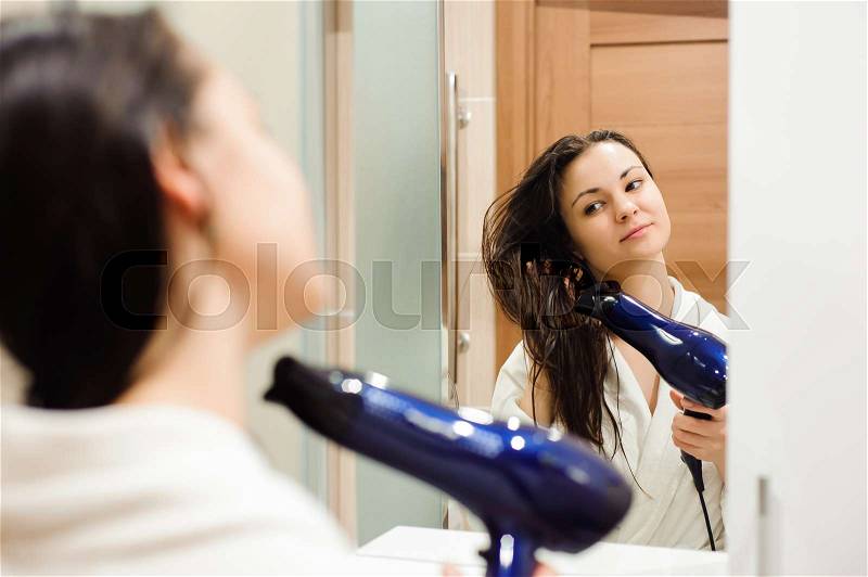 Beautiful young woman in bath towel is using a hair dryer and smiling while looking into the mirror in bathroom, stock photo