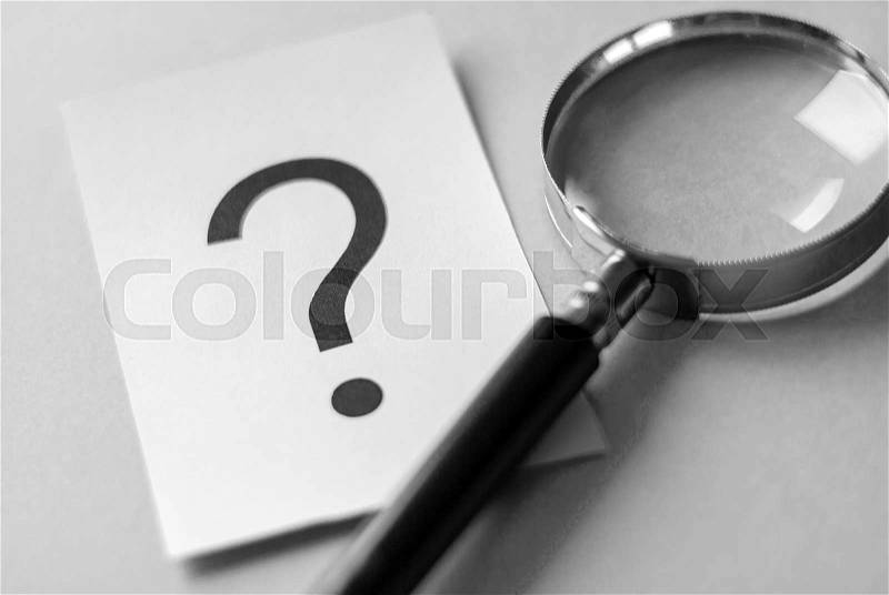 Black question mark printed on white paper card on the table next to magnifying glass, viewed in close-up. Search and questioning concept, stock photo