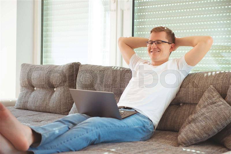 Young handsome man works at home with his laptop on couch, stock photo
