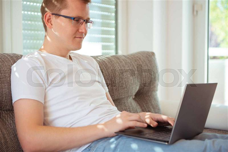 Young handsome man works at home with his laptop on couch, stock photo