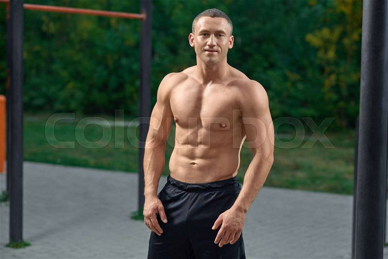 Sexy, muscular man with naked torso and biceps looking at camera, posing. Cheerful sportsman smiling, standing outdoors on street workout. Bodybuilder wearing in ..., stock photo