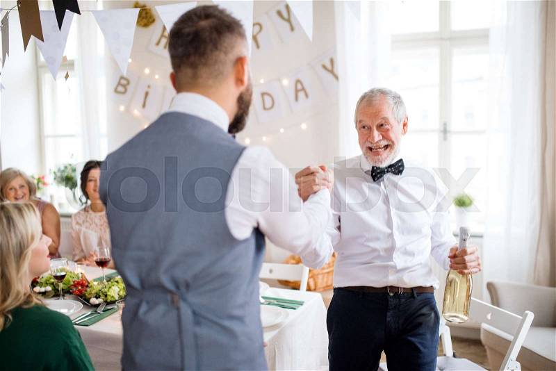 A mature man giving a bottle of wine to his father on indoor birthday party, a celebration concept, stock photo
